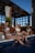 Three women in swimsuits are sitting by the edge of a pool with their feet in the water. They appear relaxed and are holding glasses of drinks. A bottle is placed nearby. The setting includes wooden structures and greenery, suggesting an outdoor pool area.