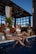 Three women in swimsuits are sitting by the edge of a pool with their feet in the water. They appear relaxed and are holding glasses of drinks. A bottle is placed nearby. The setting includes wooden structures and greenery, suggesting an outdoor pool area.