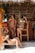 Three women in bikinis are relaxing by a rustic beach bar that appears to be made of wood and thatched roofing. Each woman is posing in a casual, relaxed manner. One is sitting on the sand and holding a drink, while the other two stand by the bar. Sunlight casts shadows on their surroundings.