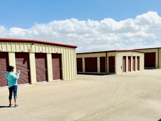 A person wearing a light blue jacket and jeans stands on a paved surface, looking at a device in their hands. The scene is set in an open area with a row of beige storage units featuring red doors. The sky is clear with scattered fluffy clouds.