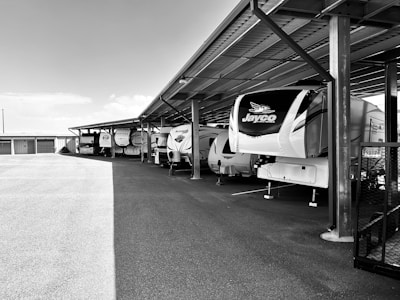 Several large camper trailers are parked under a metal carport structure. The campers are aligned in a row, with ample empty space in the foreground and additional storage units visible in the background. The structure and ground are illuminated by natural lighting, contributing to the outdoor setting.