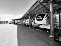 Several large camper trailers are parked under a metal carport structure. The campers are aligned in a row, with ample empty space in the foreground and additional storage units visible in the background. The structure and ground are illuminated by natural lighting, contributing to the outdoor setting.