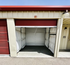 A storage unit with a partially open red roll-up door. The interior is empty, showing metallic wall panels with some insulation visible at the sides. The building has a cream color with a small label 'F-8' above the entrance.