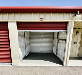 A storage unit with a partially open red roll-up door. The interior is empty, showing metallic wall panels with some insulation visible at the sides. The building has a cream color with a small label 'F-8' above the entrance.