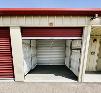 A storage unit with a partially open red roll-up door. The interior is empty, showing metallic wall panels with some insulation visible at the sides. The building has a cream color with a small label 'F-8' above the entrance.