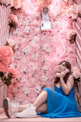 A woman in a blue dress sits on a floor covered with a lush assortment of pink and white flowers. She holds a pink vintage rotary telephone receiver to her ear, smiling slightly. Above her, a neon flamingo-shaped light illuminates the scene. The setting conveys a whimsical, playful ambiance with a soft, romantic aesthetic.