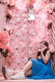 A woman in a blue dress sits on a floor covered with a lush assortment of pink and white flowers. She holds a pink vintage rotary telephone receiver to her ear, smiling slightly. Above her, a neon flamingo-shaped light illuminates the scene. The setting conveys a whimsical, playful ambiance with a soft, romantic aesthetic.