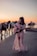 A woman in a floral dress plays a pink guitar while standing on a beach at sunset. Several horses walk along the shore in the background, adding a serene and rustic atmosphere to the scene.