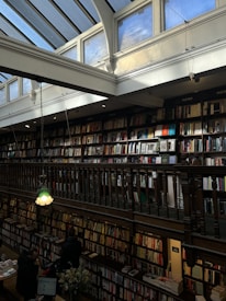 A spacious, multi-level bookstore with tall shelves filled with a variety of books. The second floor features a wooden railing overlooking the lower level. Large arched windows and a glass ceiling let in ample natural light. There are people browsing books on the first floor, along with a flower vase on a table near the entrance. Two green pendant lights hang from the ceiling.