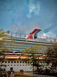 A large cruise ship named 'Carnival Horizon' is docked near a shoreline. The ship's multistory structure is visible, adorned with decks and lifeboats. Trees with lush green foliage partially obscure the lower part of the ship while a cloudy sky looms overhead.