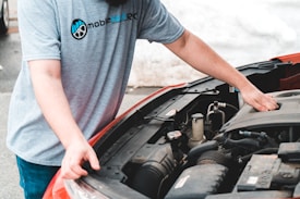 A person is working under the hood of a car, inspecting or possibly repairing the engine. The person is wearing a gray t-shirt with a logo and blue jeans. The car appears to be parked outdoors on a gray pavement surface.