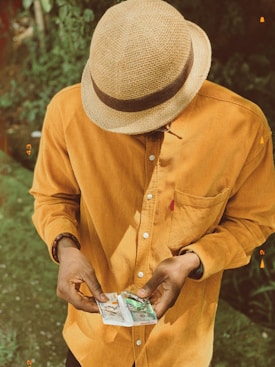 A person wearing a straw hat and a mustard-colored shirt is looking down at a handful of money. The setting appears to be outdoors, with greenery in the background.
