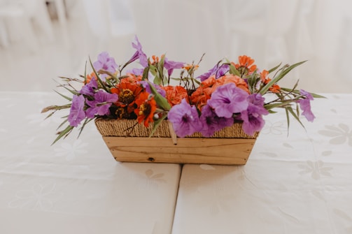 A rectangular, woven wooden basket filled with a vibrant arrangement of purple and orange flowers. The basket rests on a light-colored tablecloth with subtle floral patterns.