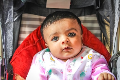 A baby is looking directly at the camera while sitting in a stroller with a red padded interior. The baby is wearing a light-colored outfit with colorful patterns, including small text and illustrations. The background includes striped fabric and the edges of the stroller frame.