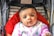 A baby is looking directly at the camera while sitting in a stroller with a red padded interior. The baby is wearing a light-colored outfit with colorful patterns, including small text and illustrations. The background includes striped fabric and the edges of the stroller frame.