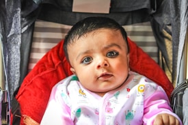 A baby is looking directly at the camera while sitting in a stroller with a red padded interior. The baby is wearing a light-colored outfit with colorful patterns, including small text and illustrations. The background includes striped fabric and the edges of the stroller frame.