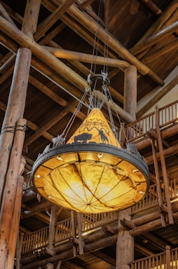 A large rustic chandelier hangs from a high wooden ceiling. The chandelier features an ornate design with silhouettes of animals and mountains, emitting a warm, ambient glow. The surrounding architecture is constructed with sturdy wooden beams and railings, adding to the lodge-like atmosphere.