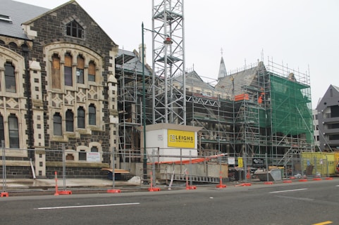 A construction site featuring an old stone building partially covered in scaffolding and green safety netting. The building has Gothic architectural elements with arched windows and stone detailing. In the foreground, there are barriers and construction signage, indicating ongoing restoration work.