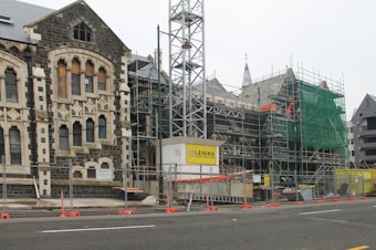 A construction site featuring an old stone building partially covered in scaffolding and green safety netting. The building has Gothic architectural elements with arched windows and stone detailing. In the foreground, there are barriers and construction signage, indicating ongoing restoration work.