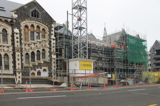 A construction site featuring an old stone building partially covered in scaffolding and green safety netting. The building has Gothic architectural elements with arched windows and stone detailing. In the foreground, there are barriers and construction signage, indicating ongoing restoration work.