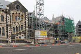 A construction site featuring an old stone building partially covered in scaffolding and green safety netting. The building has Gothic architectural elements with arched windows and stone detailing. In the foreground, there are barriers and construction signage, indicating ongoing restoration work.