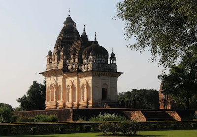 A historical temple with intricate architectural details stands amidst lush greenery. The temple features multiple domes with ornate carvings, and its faded stone surface adds to its ancient charm. Tall trees and well-manicured gardens surround the structure, enhancing its majestic presence.