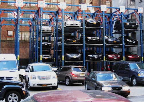 A multi-level car parking structure with various vehicles stacked on top of each other in a vertical arrangement. The structure is made of metal beams and platforms, painted blue and red, and is situated in an urban setting with brick buildings in the background. Several cars are parked at ground level in front of the structure.
