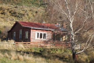 A rustic wooden house with a red roof is nestled in a rural landscape. The house is surrounded by dry grass and bare trees, suggesting a cold season. The wooden fence enhances the countryside ambiance, while the sloping grassy hills in the background add depth to the setting.