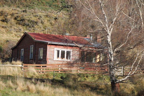 A rustic wooden house with a red roof is nestled in a rural landscape. The house is surrounded by dry grass and bare trees, suggesting a cold season. The wooden fence enhances the countryside ambiance, while the sloping grassy hills in the background add depth to the setting.