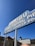 A vintage-style sign advertising Malibu Pier, sport fishing, live bait, and charter services. The sign has a retro font with white lettering against a blue background, supported by two white poles. It is set against a clear blue sky with some buildings partially visible in the background.