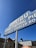 A vintage-style sign advertising Malibu Pier, sport fishing, live bait, and charter services. The sign has a retro font with white lettering against a blue background, supported by two white poles. It is set against a clear blue sky with some buildings partially visible in the background.