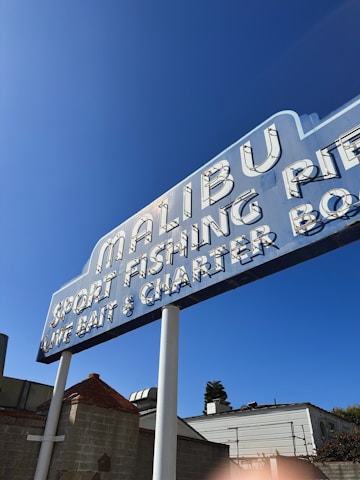 A vintage-style sign advertising Malibu Pier, sport fishing, live bait, and charter services. The sign has a retro font with white lettering against a blue background, supported by two white poles. It is set against a clear blue sky with some buildings partially visible in the background.