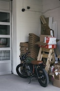 A motorcycle is parked inside a storage room or garage, alongside stacks of cardboard boxes and packaged materials. The concrete floors, walls constructed of cinder blocks, and presence of security cameras give an industrial feel. In the background there are more stacked objects, including wooden planks and panels, and the garage door is partially visible.