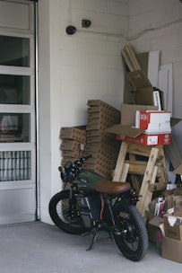 A motorcycle is parked inside a storage room or garage, alongside stacks of cardboard boxes and packaged materials. The concrete floors, walls constructed of cinder blocks, and presence of security cameras give an industrial feel. In the background there are more stacked objects, including wooden planks and panels, and the garage door is partially visible.