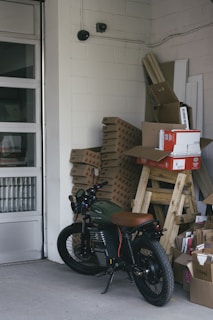A motorcycle is parked inside a storage room or garage, alongside stacks of cardboard boxes and packaged materials. The concrete floors, walls constructed of cinder blocks, and presence of security cameras give an industrial feel. In the background there are more stacked objects, including wooden planks and panels, and the garage door is partially visible.