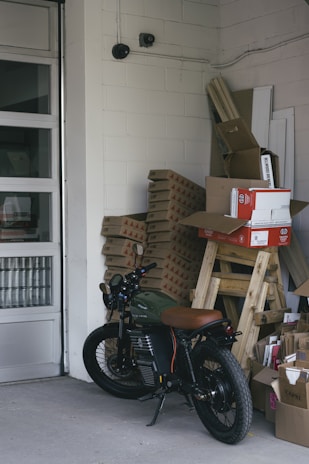 A motorcycle is parked inside a storage room or garage, alongside stacks of cardboard boxes and packaged materials. The concrete floors, walls constructed of cinder blocks, and presence of security cameras give an industrial feel. In the background there are more stacked objects, including wooden planks and panels, and the garage door is partially visible.