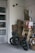 A motorcycle is parked inside a storage room or garage, alongside stacks of cardboard boxes and packaged materials. The concrete floors, walls constructed of cinder blocks, and presence of security cameras give an industrial feel. In the background there are more stacked objects, including wooden planks and panels, and the garage door is partially visible.