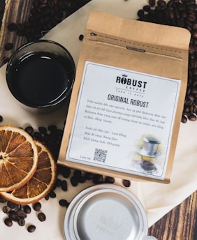 A package of Robust coffee sits on a surface surrounded by coffee beans. Next to the package, there is a cup filled with black coffee and several slices of dried orange. The package features labeling and text about the coffee, and a QR code is visible. A silver lid or cup is partially seen at the bottom.