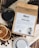 A package of Robust coffee sits on a surface surrounded by coffee beans. Next to the package, there is a cup filled with black coffee and several slices of dried orange. The package features labeling and text about the coffee, and a QR code is visible. A silver lid or cup is partially seen at the bottom.