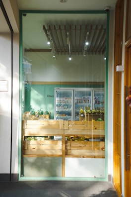 A small, well-lit shop displays wooden crates filled with various fruits and vegetables. The crates are neatly arranged in front of a glass wall, and beyond them, a refrigerated section can be seen with more produce. The ceiling features modern lighting fixtures, and the walls are painted in a light green color.