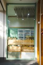 A small, well-lit shop displays wooden crates filled with various fruits and vegetables. The crates are neatly arranged in front of a glass wall, and beyond them, a refrigerated section can be seen with more produce. The ceiling features modern lighting fixtures, and the walls are painted in a light green color.