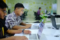 Several individuals are focused on their laptops in a modern office environment. The space features glass walls with decorative patterns, and various office supplies are visible on the desks.