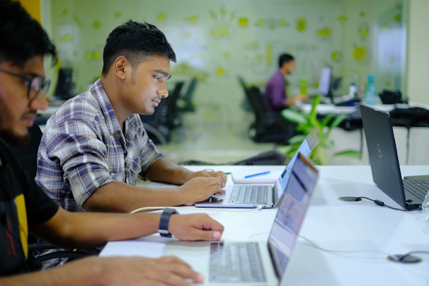 Several individuals are focused on their laptops in a modern office environment. The space features glass walls with decorative patterns, and various office supplies are visible on the desks.