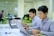 Two individuals are focused on their laptops in an office setting. The environment is modern with a bright and clean aesthetic. Green plants are placed around the room, and in the background, another person is visible working in an open space with glass partitions.