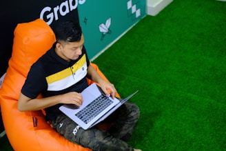 A person is seated on an orange bean bag chair while working on a laptop. The environment appears casual and modern, with a green floor that resembles artificial grass. A wall with some geometric designs and text can be seen as part of the background.
