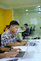People are working on laptops at a modern office setup. The environment appears collaborative and tech-focused, with laptops, notebooks, and a bright, colorful interior featuring orange and green tones.