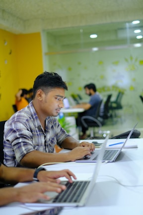 People are working on laptops at a modern office setup. The environment appears collaborative and tech-focused, with laptops, notebooks, and a bright, colorful interior featuring orange and green tones.