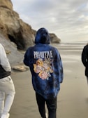 A person in a hoodie with floral designs walks along a beach. The text 'PRIMITIVE' is printed on the back. The sandy beach is bordered by rocky cliffs, and another person is partially visible. The sky is overcast, casting a calm atmosphere.