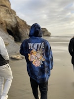 A person in a hoodie with floral designs walks along a beach. The text 'PRIMITIVE' is printed on the back. The sandy beach is bordered by rocky cliffs, and another person is partially visible. The sky is overcast, casting a calm atmosphere.