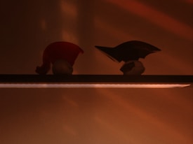 Silhouettes of two small, abstract objects are placed on a shelf, illuminated by a warm light creating a sharp contrast against the background. The left object resembles a red hat or cap, while the right appears to have a curved design, possibly aerodynamic. The lighting creates a moody and contemplative atmosphere.